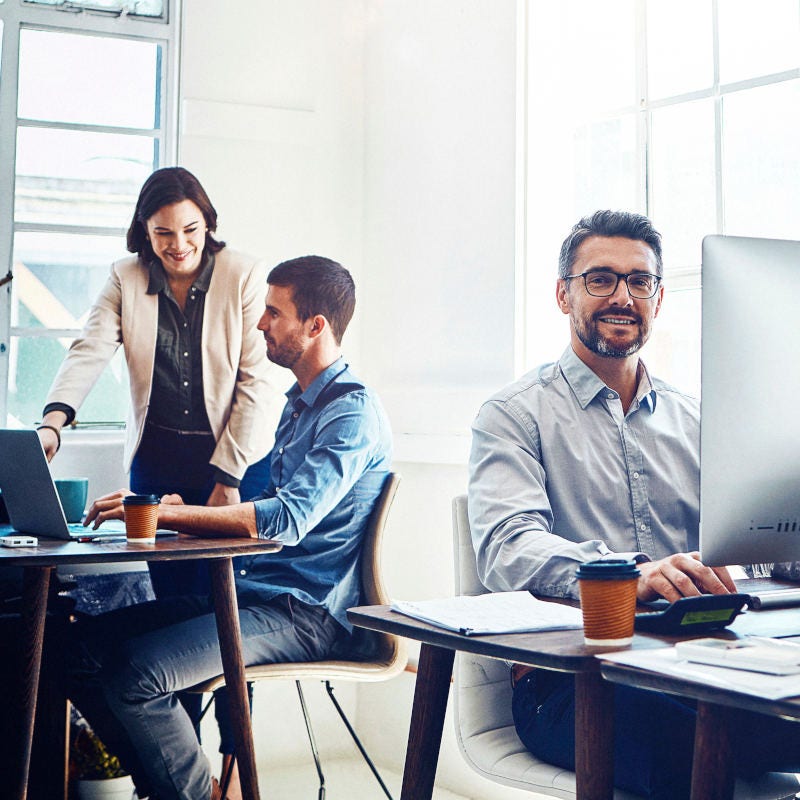 Hombre sonriendo en su escritorio trabajando en computador. Tras él se encuentran dos compañeros conversando Hombre sonriendo en su escritorio trabajando en computador. Tras él se encuentran dos compañeros conversando
